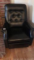 Front view of the black pleather rocker recliner chair showing button tufted backrest and padded armrests on hardwood floor near brick wall.