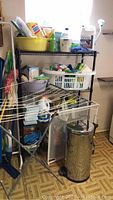 Overview photo showing metal shelving unit with multiple cleaning supplies, laundry baskets, drying rack, and metal waste bin on floor