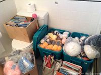 Photo showing two Rubbermaid containers packed with various yarn skeins and balls along with boxes of yarn on a bathroom floor.