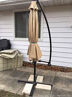 Closed beige offset patio umbrella attached to black metal stand with four patio stone weights at the base. Photos taken outdoors against a house wall with patio flooring visible.
