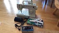 Wide view showing Singer Debutante 7021 sewing machine with attached cord, black foot pedal next to it, boxed Kenmore buttonhole attachment and packaged white-handled Singer scissors