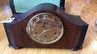 Front view of wooden mantle clock showing curved top and glass front face with gold Arabic numerals and brass bezel.
