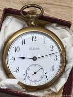 Front view of the gold-filled pocket watch with white dial, black Arabic numerals, blue hands, and brand name D.E. Black Calgary.