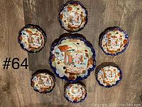 Seven Japanese ceramic bowls: one large scalloped bowl in the center surrounded by six smaller bowls, all with detailed painted geisha scenes and vibrant colors including orange, blue, green, gold, and white