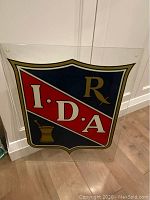 Front view of the glass sign showing the clear, colorful IDA logo with red, navy blue, white, gold, and black accents.