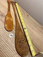 Two miniature wooden paddles placed on a wood surface with tape measure showing sizes (approx. 12 inches and 17 inches).