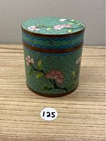 Closed view of cylindrical cloisonne box with floral design on turquoise background, copper bands, and flat lid.