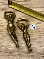 Two brass female figure bottle openers placed next to measuring tape showing lengths approximately 5 and 4 inches.