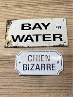 Top view of the two metal signs 'BAY WATER' and 'CHIEN BIZARRE' placed on a wooden surface.