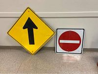 Two road signs leaned against a wall: yellow diamond sign with black arrow and square white sign with red no entry symbol.