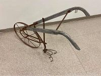Full view of antique farm tiller showing wooden handles, rusted metal frame and tines, and metal spoked wheel on a floor.