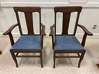 Front view of two oak dining arm chairs with blue cushioned seats, vertical slatted backs, and curved armrests.