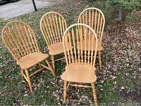 Four solid oak arrowback chairs arranged outdoors on grass with fallen leaves, showing wood grain, seat design, backrest spindles, and turned legs.