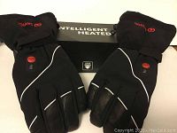 Pair of black heated gloves with reflective piping and red power button laid side by side on white surface.