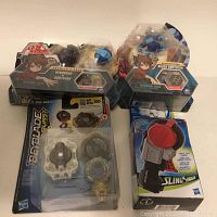 Four packages including Beyblade Burst toy top set, two Bakugan Siege Gorthion figures, and SlingShock launcher in box. Packaging shows some damage.