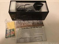 Photo of the boxed tactical rifle scope with foam padding and instruction sheet underneath, includes cleaning cloth and batteries pack.