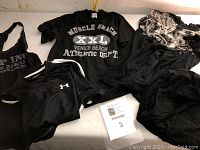 Five black men's T-shirts laid out on a flat surface, includes Muscle Beach XXL graphic T-shirt, Under Armour branded athletic shirt with white sleeve stripes, and three other black T-shirts of varying styles.