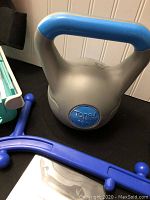 Close-up of Tonex branded silver 10 lb kettlebell with blue handle and a blue flexible back massager on a table