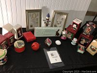Overview of assorted holiday decorations displayed on black cloth including boxed ornaments, decorative figurines, and framed art