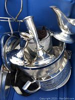 Shows a silver plated gravy boat with attached base, a handled bucket, a pair of tapering candlesticks, round trays underneath, and an oval serving dish at bottom