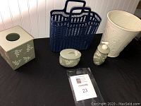 Bathroom accessory assortment arranged on black surface, showing cream wastebasket, navy blue basket, ceramic soap dispenser and lidded jar, and tissue box cover.
