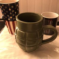 Three ceramic coffee mugs displayed: one green grenade-shaped mug front and center, a tall American flag patterned mug at left, and a smaller black mug with an American flag design at right.