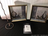 Two framed bathroom-themed artwork prints and a metal toilet tissue holder displayed on a black surface against a beadboard wall.
