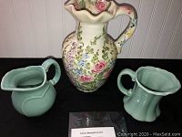 Three vintage pottery pitchers displayed against a white paneled wall and black surface: one large floral and two small green pitchers.