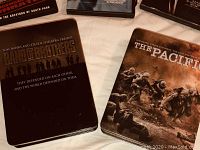 Band of Brothers and The Pacific DVD boxed sets lying side by side on a beige surface. Both are collector's edition cases with detailed cover art depicting soldiers.