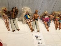 Seven Barbie and Ken dolls arranged sitting against a light wood background, showcasing outfits and hair details