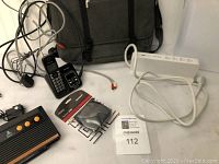 Overall photo showing Atari console, phone set with headset, white Belkin power strip, and black computer bag in background.