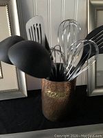 Copper utensil holder with black kitchen utensils inside including spoons, spatulas, and whisks with framed floral artwork in background.