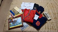 Photo of assorted Alma College apparel including sweaters, polo shirts, visor, and small memorabilia items on a beige patterned cloth.