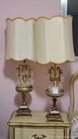 Front view of the pair of table lamps showing average size, base design, crystal pendants, and scalloped cream shades with gold trim.