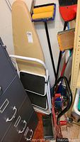 Photo showing ironing board, step stool, brooms and dustpans in storage area