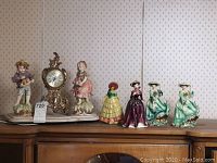 Overview of four small ceramic lady figurines and mantle clock flanked by two bisque figurines on wooden cabinet.