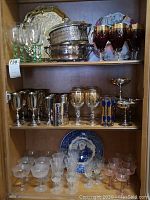 Full view of cabinet with shelves displaying glassware, silver plated tray, vegetable dish, and decorative plates.