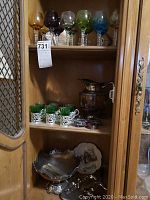 Cabinet shelf with colored stemmed glasses in various colors on the top shelf, silver plated pitcher, salters, candy dish, green glass shooters in silver holders on the middle shelf, silver plated centerpiece vase bowl, commemorative plate, and chrome silent butler on lower shelf.