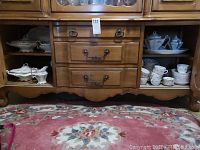 Wooden cabinet with shelves holding multiple pieces of white china with gold rims and green floral laurel patterns. Includes plates, cups, and serving items.