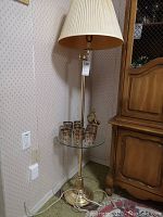 Full view of the brass floor lamp with glass table and pleated lamp shade, standing next to a wooden cabinet