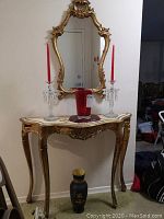 Full view of demi lune hall table with decorative gold finish and Queen Anne legs, with two glass candlesticks holding red candles, ruby glass vase on wooden platter on top, black ceramic vase on floor in front.