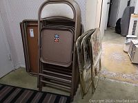 Stack of 4 brown metal folding chairs with padded seats and backs next to 3 decorative folding TV trays showing floral patterns and a black folding card table.