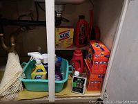 Shelf under sink with assorted cleaning products including Tide detergent boxes, Drano bottle, SOS steel wool pads, Lysol spray, and other cleaning bottles in a green plastic basket.