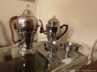 Photo showing two polished stainless steel coffee percolators, one with a spigot and the other with a pour spout, alongside a cream and sugar set on a tray.
