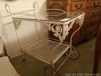Full view of off-white metal framed tea cart with glass top, floral detailing, large and small wheels, and mesh bottom shelf.
