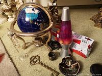 Brass globe showing blue and gold tones next to purple lava lamp and original box, handcrafted glazed clay cup and ashtray, thermometer, and brass key holder on carpeted floor.