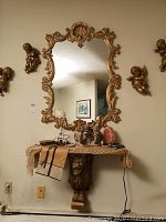 Photo shows gold toned ornate shell and scroll framed wall mirror above wall mounted lion head table with decorative items on table including salt lamp and cherubs