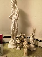 Wide shot showing all items together on a floor near a wall: tall Grecian statue, cherub pedestal base, ceramic pitchers, and basket carrying figurines.