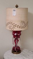 Full view of ruby glass table lamp with beige leaf-patterned shade and brass finial on top