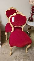 Pair of red velvet upholstered chairs with ornate carved light painted wood frames, featuring floral designs on backrest, visible from front angle.